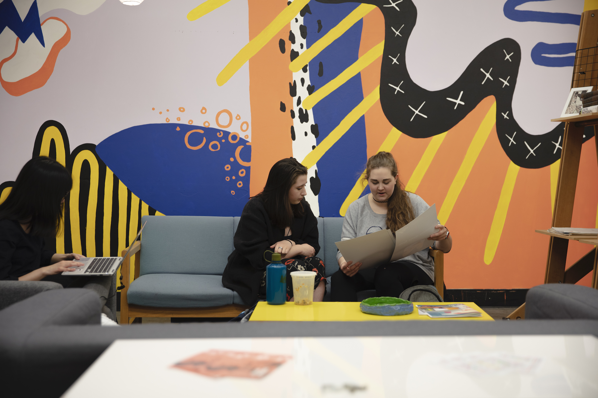 Two women studying on a sofa in front of a colorful mural.