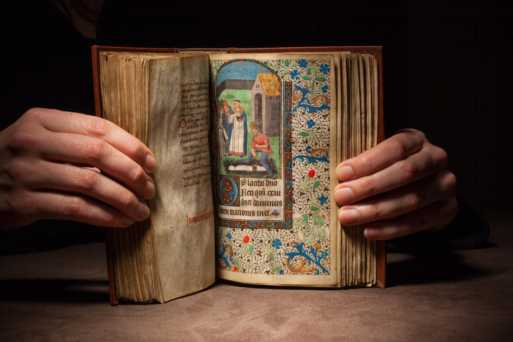 Hands holding open a medieval book of your to a page with illustration of a burial.