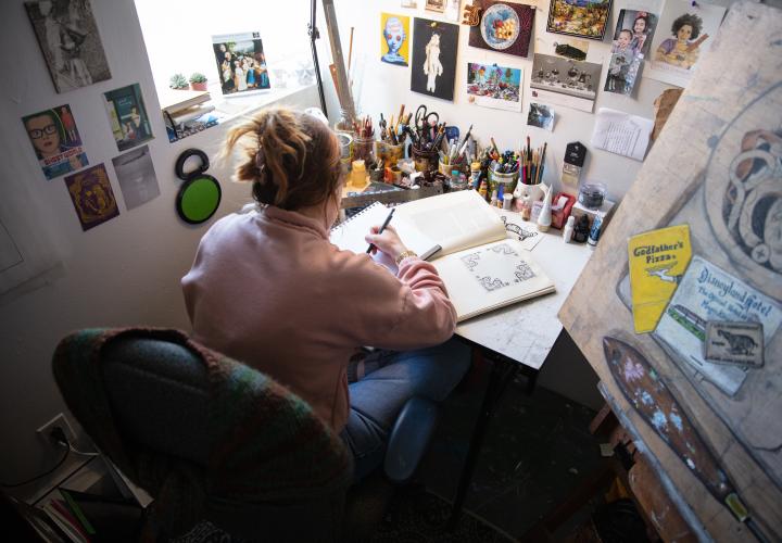 Graduate student working at a drawing table by a window, surrounded by reference images and art supplies