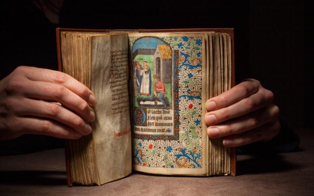 Hands holding open a medieval book of your to a page with illustration of a burial.