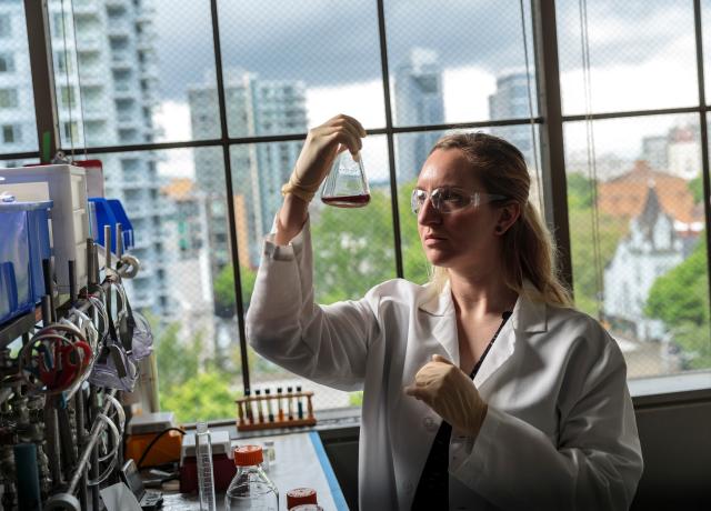 Student scientist looks at a flask with a dark colored liquid inside. Portland cityscape can be seen through the windows in the background.