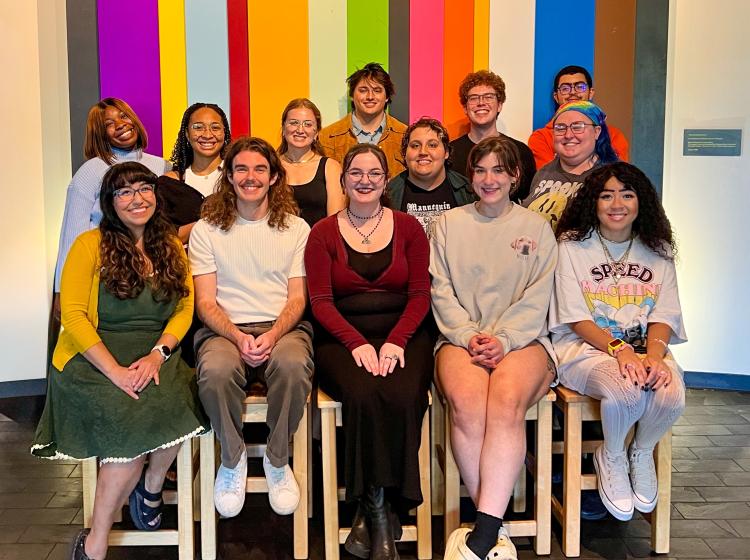 Photo of current graduate students in 3 rows with 5 people sitting on stools before standing rows. The background is a wall painted with colorful stripes.