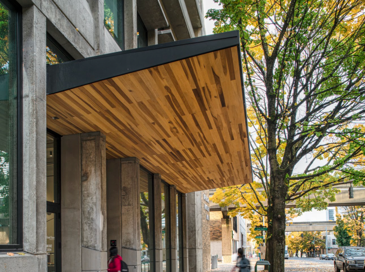 One student stands on the sidewalk looking into the concrete building while another stands underneath a wood awning