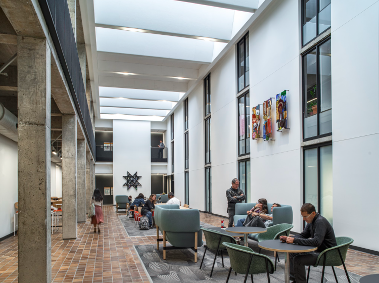 Students sit at tables and couches located in a long, open space decorated by colorful art and windows looking into offices