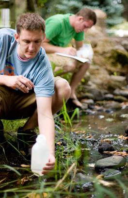 Male student taking a water sample from a stream. Another male student is in the background making notes.