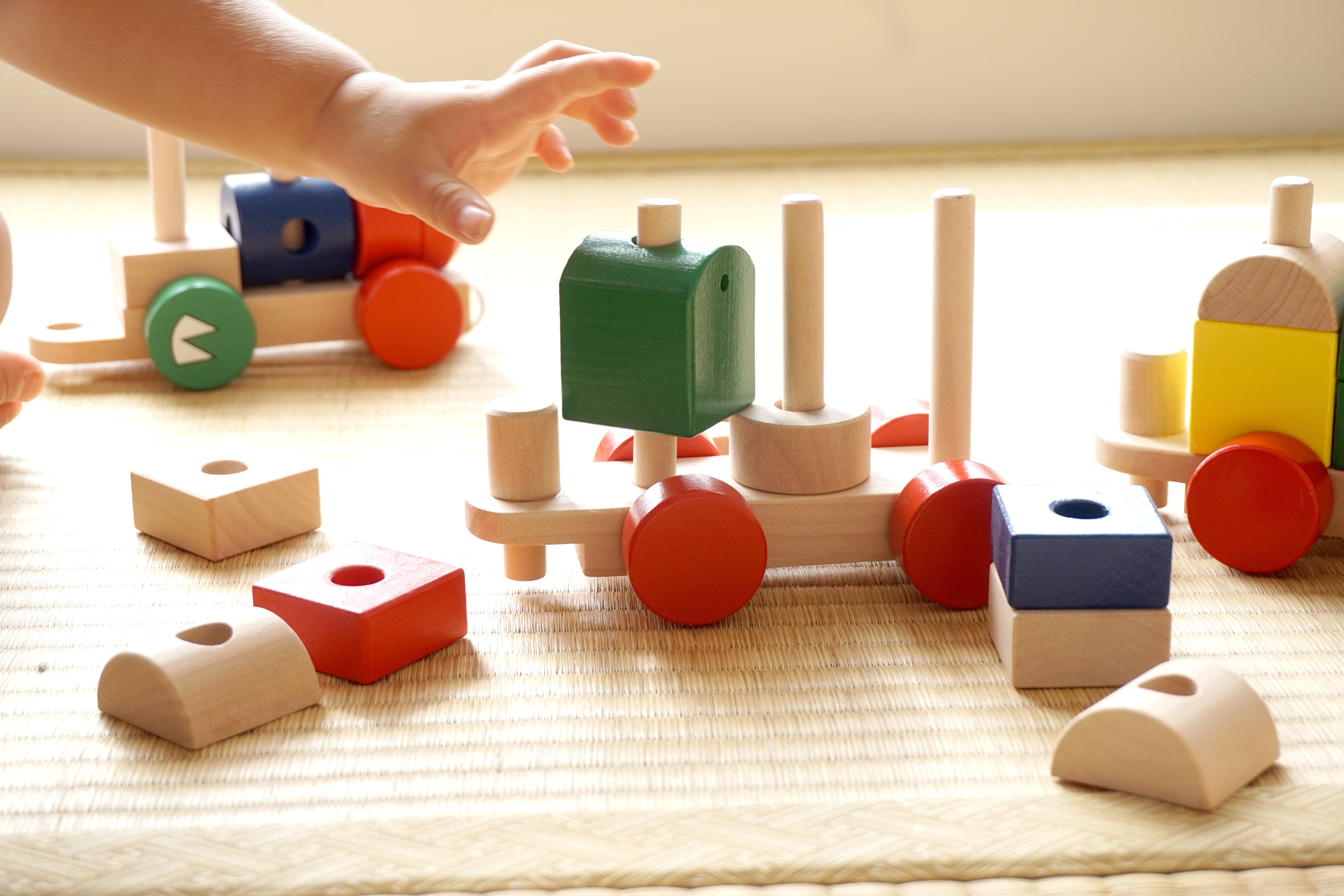 A child's hand reaches for a bright green wooden block surrounded by a colorful wooden train set.