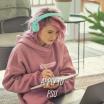 A young woman with pink hair and hoodie sits before a laptop, writing in a notebook and wearing mint-colored headphones.