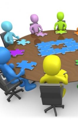 individuals sitting around a round table at a meeting with a puzzle in the center of the table. Each individual has in front of them a puzzle piece. 