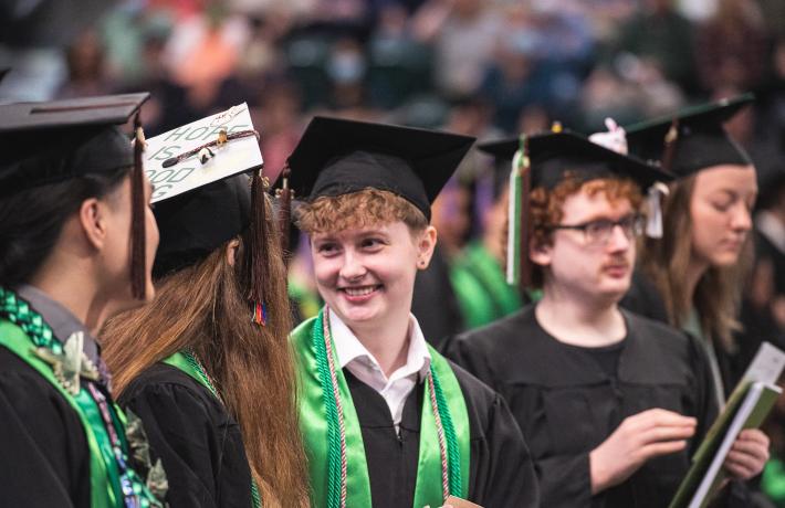 Three students in graduation caps and gowns with green regalia stand in a crowd. Student with red hair smiles at another student with long brown hair.
