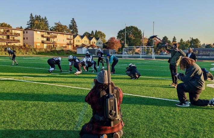 Football team sets up for a play while student in the foreground films the action