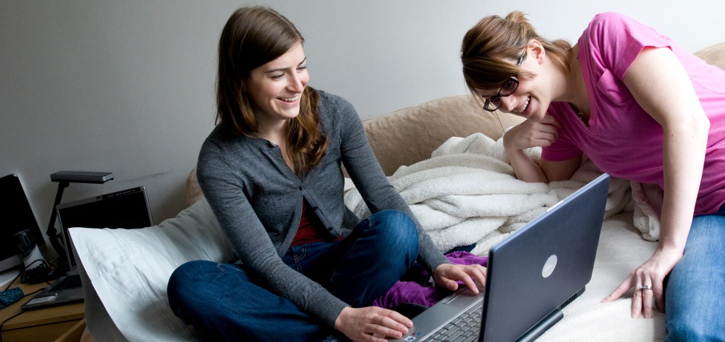 Two students sitting on a bed and one has a laptop pulled out, while the other is leaning over to look at the screen