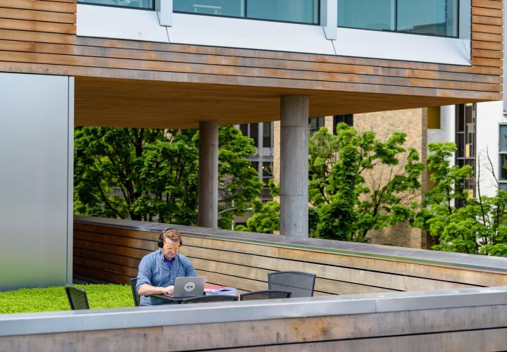 Photo of a student sitting outside on the green roof of the Karl Miller Center on his laptop and headphones doing homework all alone.