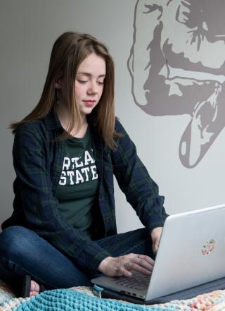 PSU student sitting on a bed in a dorm room and working on a laptop