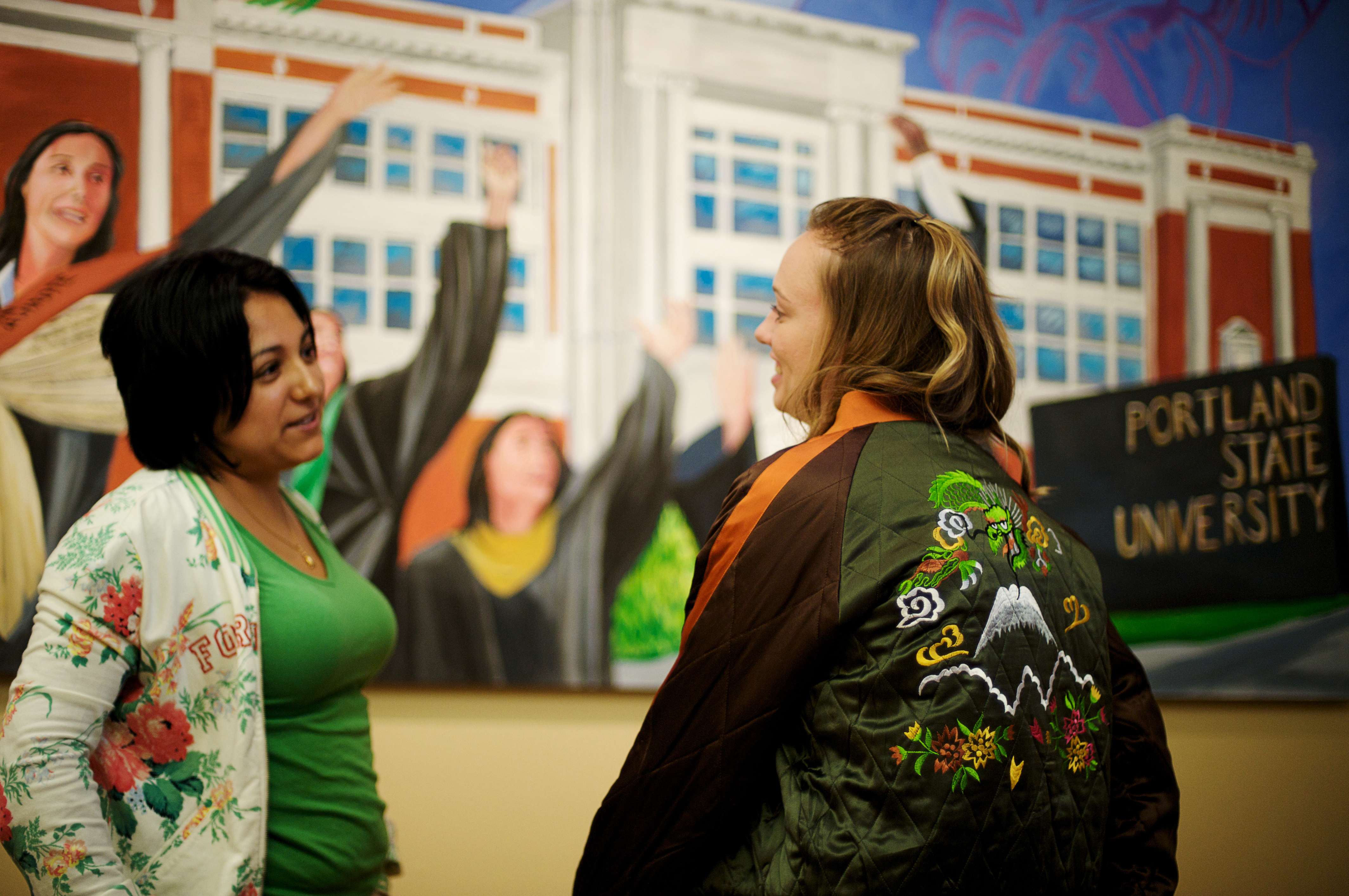 Two students standing and having a conversation in front of a mural depicting PSU graduates in caps and gowns.