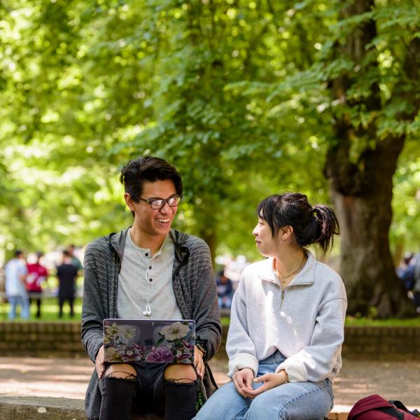 Two students sitting in front of many leafy green trees. One has a laptop.