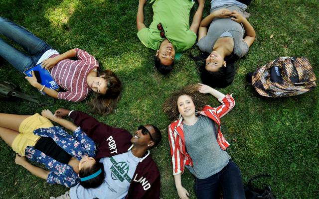 Group of college students with different ethnicities laying on their backs on top of green grass.