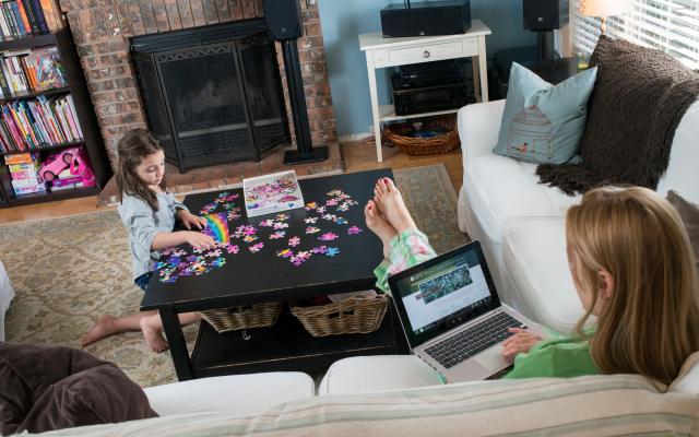 Mom on couch with laptop, daughter playing with puzzle at coffee table