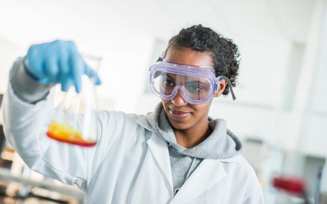 Student wearing labcoat and safety goggles holding a beaker with a yellow liquid