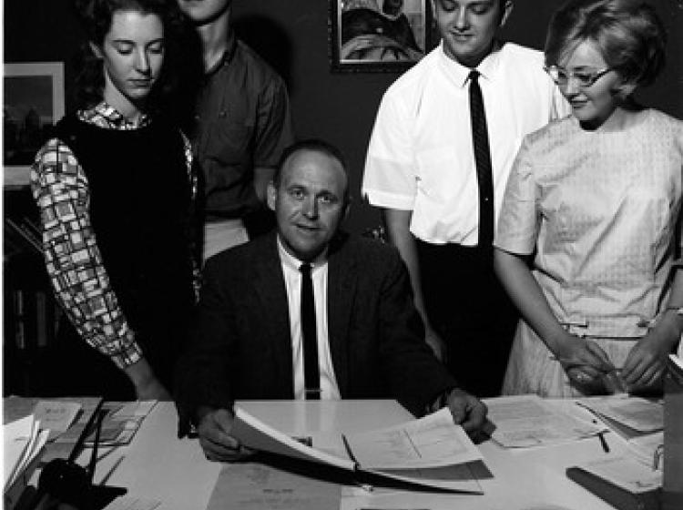 A black and white scene of a man in a suit and tie sits desk surrounded by four students all in 1960s attire.