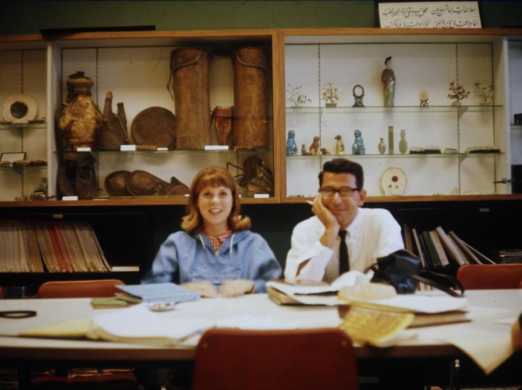 A woman and a man are sitting at a table covered in books and papers with historical artifacts on display in the background.