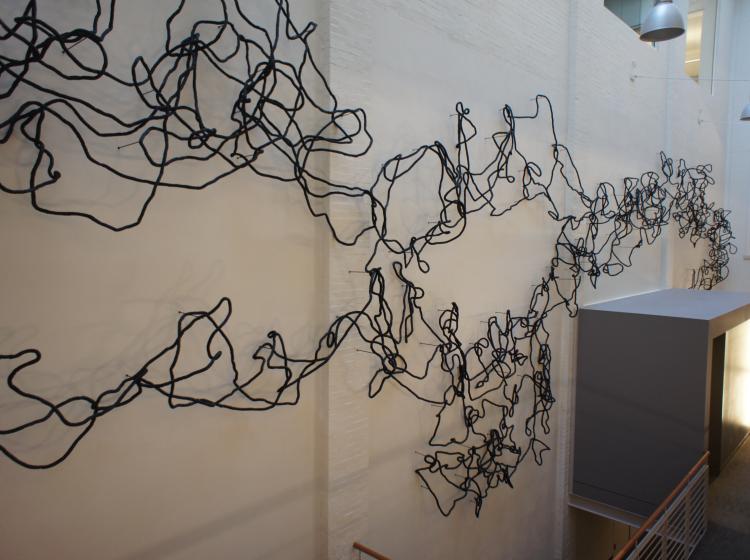 A view of the artwork looking slightly down into the atrium from above. A large amount of black rope-like cord arcs and loops along the wall, like scribbled lines