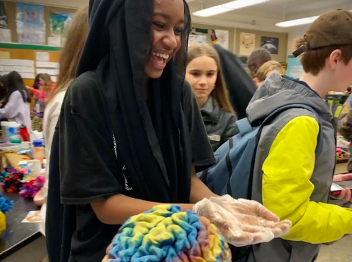 In the foreground, a brain painted in rainbow colors. Behind it, a grinning girl holds out her hands. Photo courtesy of NW Noggin.