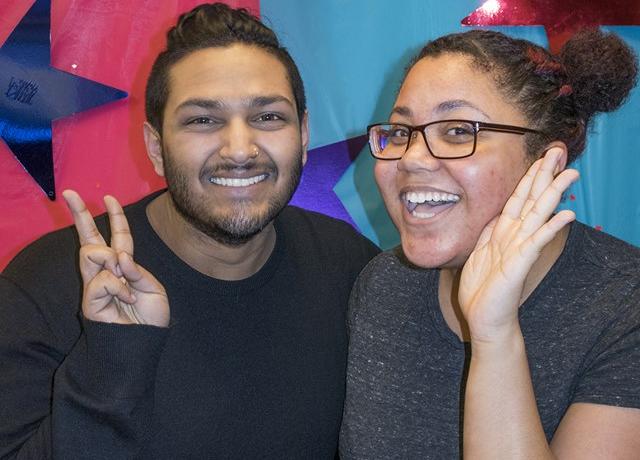 two black and brown queer people smile enthusiastically in front of a pink and blue starry background flashing peace signs