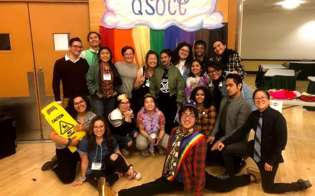 a group of QTBIPOC smile in front of a rainbow QSOCC sign. one is doing the splits, one clutches a wet floor sign jokingly. the mood is silly. 