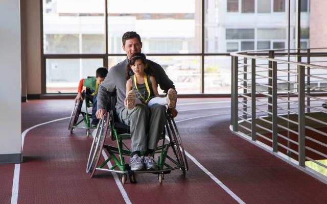 A youth member smiles and rides on the lap of a caregiver in a sports chair on the indoor track