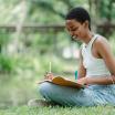 A woman sits crossed-legged in a park on the grass smiling and writing or drawing in a notebook.
