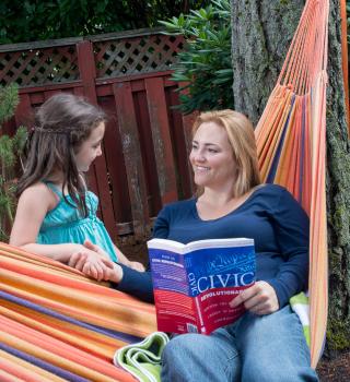 a child stands next to a hammock where a woman is sitting and reading a text book