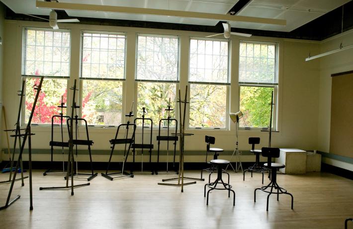 Empty class studio with metal easels and chairs scattered in the room while the room is illuminated by the sunlight from the windows behind them.