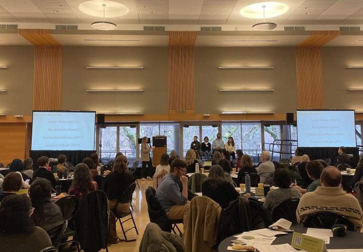 Symposium attendees gathered at tables watching four student speakers sitting on a small stage
