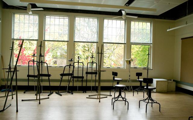 Empty class studio with metal easels and chairs scattered in the room while the room is illuminated by the sunlight from the windows behind them.
