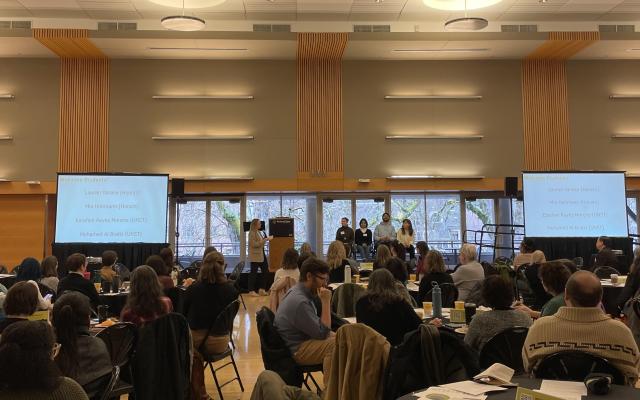 Symposium attendees gathered at tables watching four student speakers sitting on a small stage