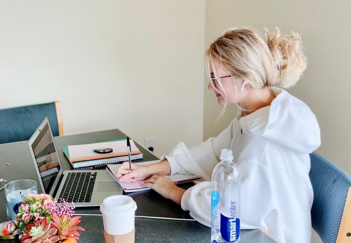 A female student with blond hair and glasses writes in a notebook while looking at a laptop.