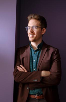 Man with glasses and sandy hair and goatee wearing a blazer and crossing his arms smiles at camera.