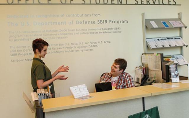 Two people, one standing and one sitting, behind a reception desk