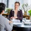 A sandy haired woman wearing a blazer listens to another person across a desk.