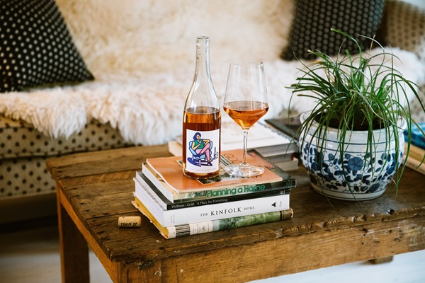 A bottle of rose next to a glass of rose sitting on a stack of books on a coffee table of a living room.