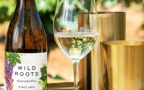 A bottle of Wild Roots Wine next to a glass of wine on a table in front of a vineyard.