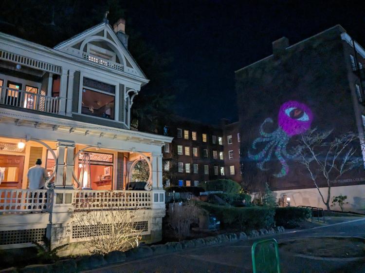 Simon Benson House lit up at night with a projected purple octopus with an eyeball on it lit up on the side of the Montgomery dorm brick wall.