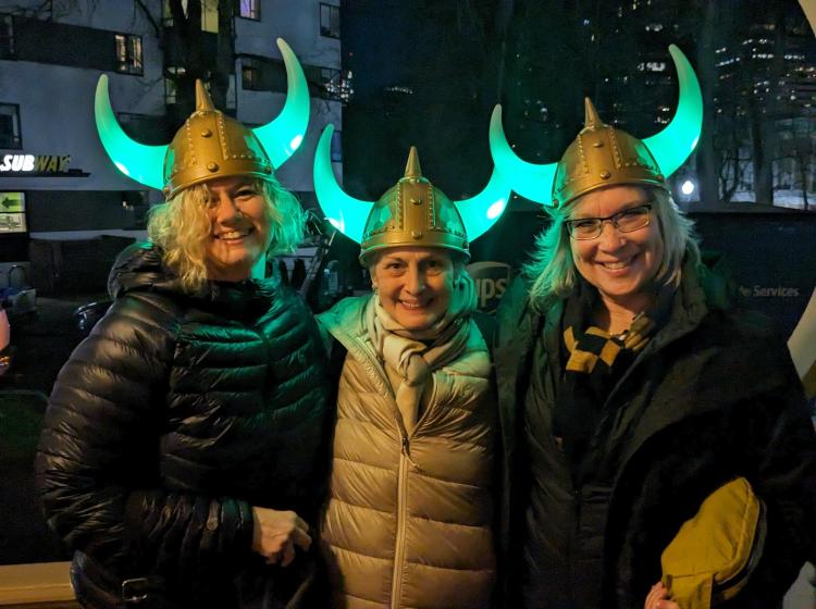 Three women wearing light-up Viking helmets with glowing green horns in the evening at the Winter Light Festival.