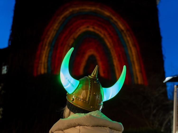 Back of a person's head wearing a light-up viking helmet looking at a rainbow projected onto the building in front of them.