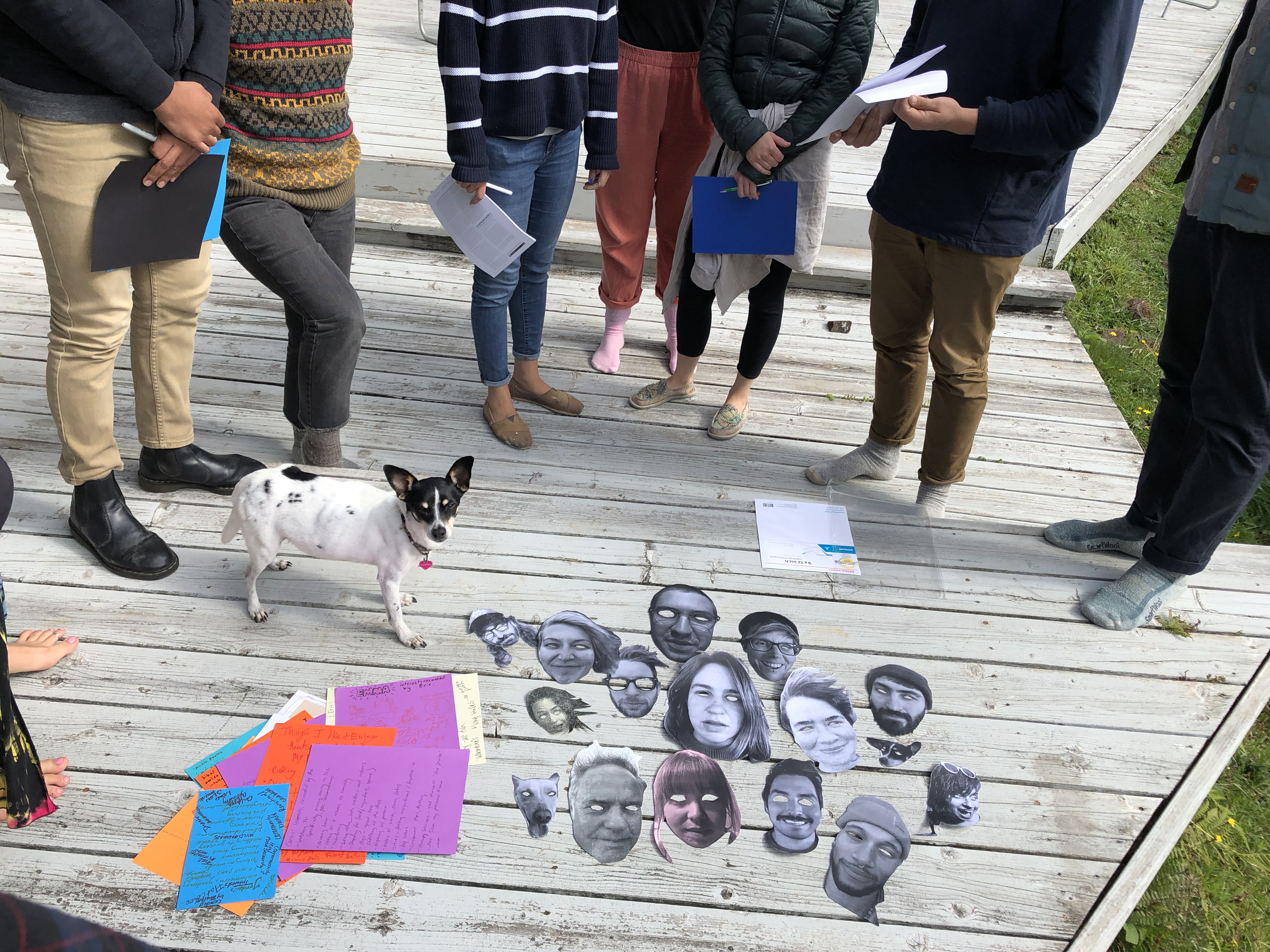 An assortment of masks laid out on a wooden deck before a group of students and a small dog