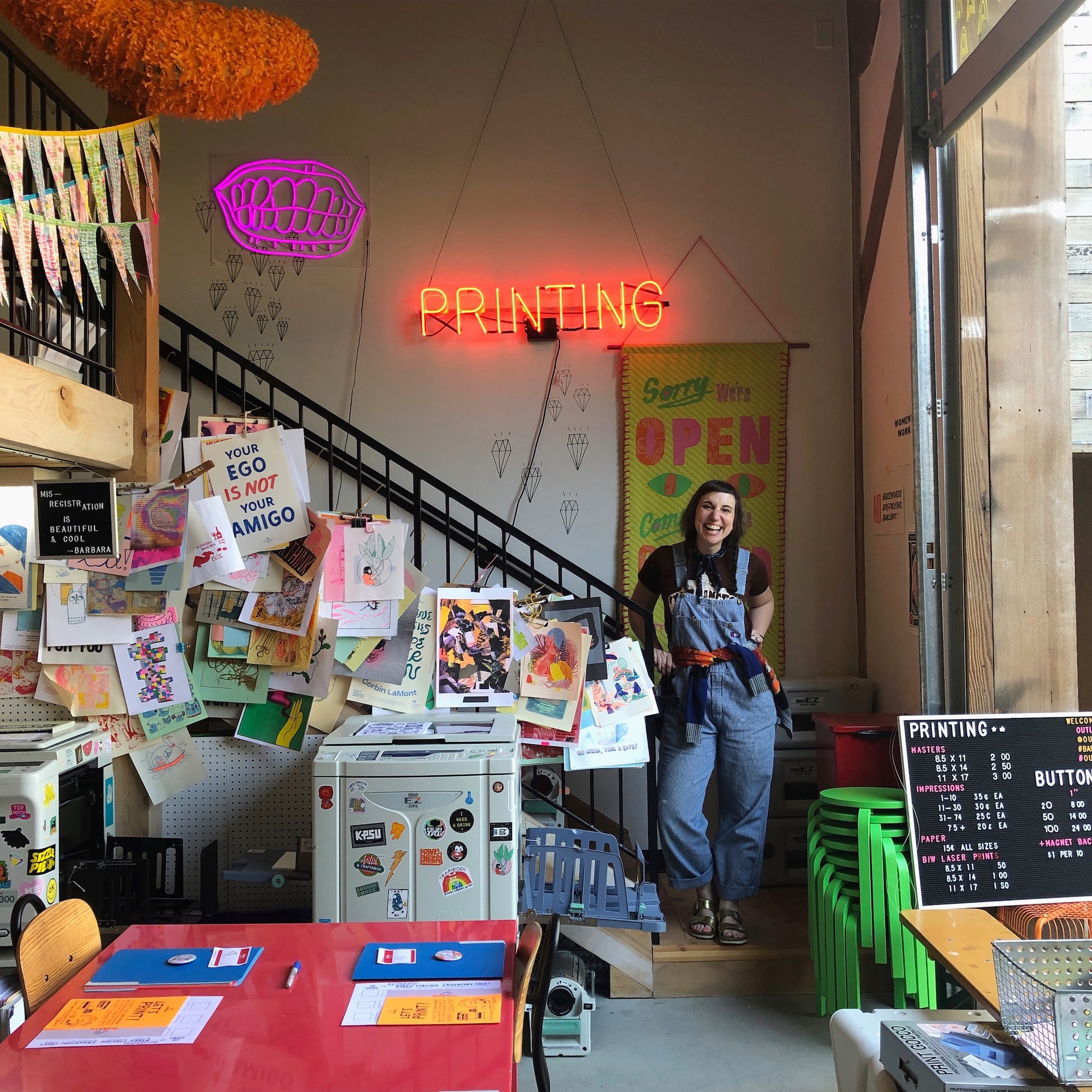 Kate Bingaman Burt stands next to a Risograph printer under a neon sign that says "Printing"