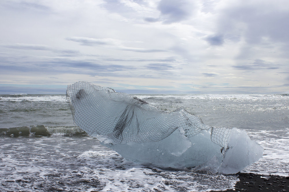 Large chunk of ice on a beach draped in a textile artwork net by Sarah Nance