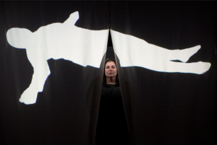 Sabina Haque seen behind a parted curtain with a white silhouette on black of a prone figure.