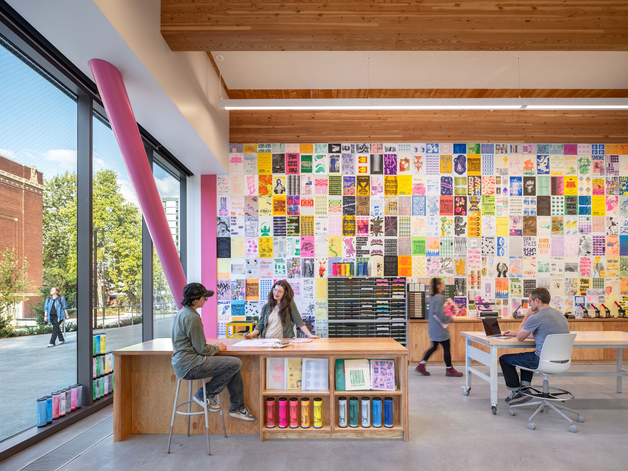 Students work in ABX 150, the Publication Studio, a room featuring a floor to ceiling wall of colorful student-designed posters and Risograph printing equipment.