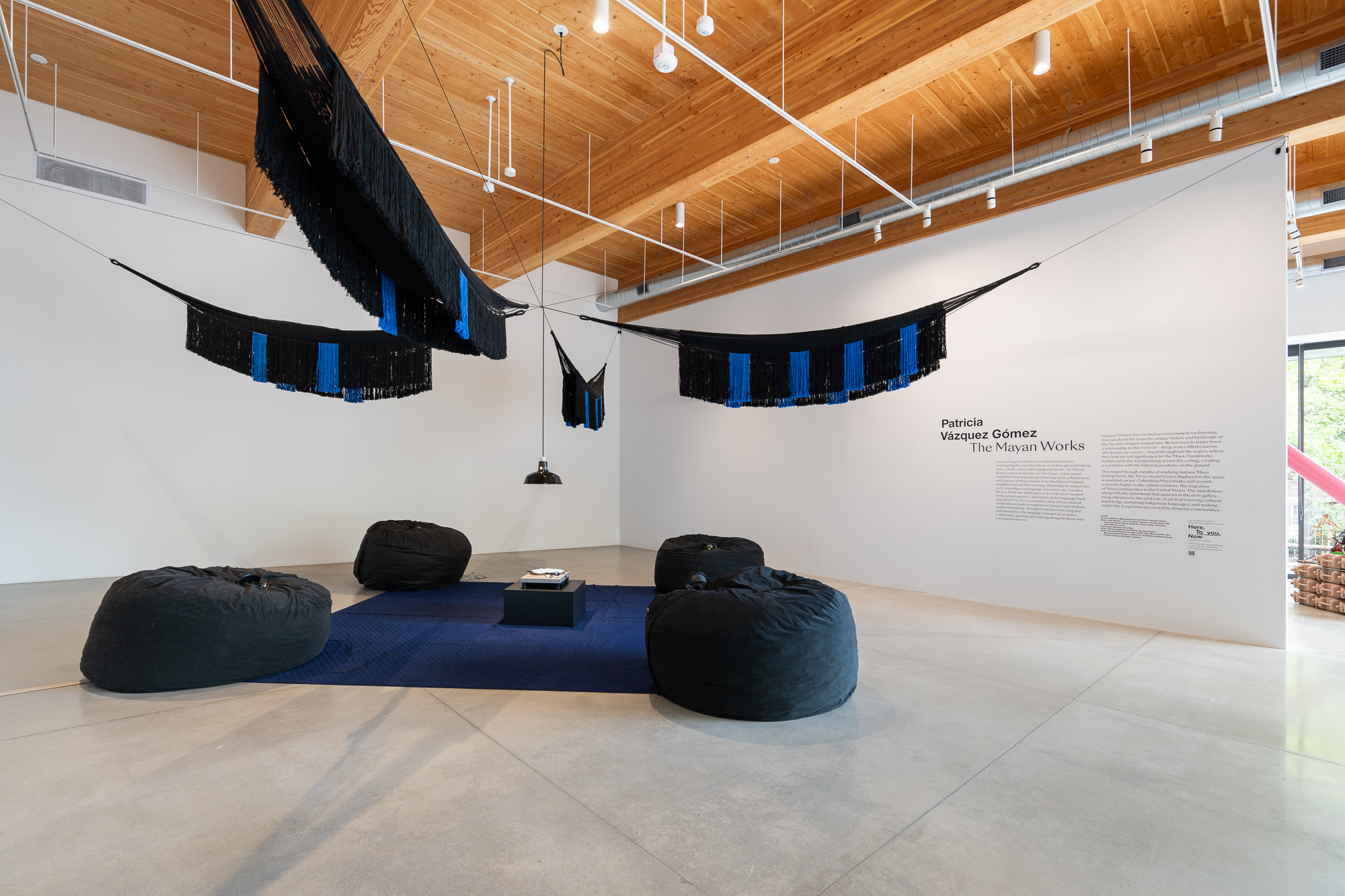 Exhibition with 4 black and blue hammocks hung high above a blue carpet. At the center of the carpet is a turntable and a pendant lamp. 4 black pillow chairs with headphones are at each corner of the carpet.